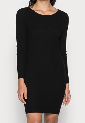 Frau trägt ein eng anliegendes, langärmeliges schwarzes Strickkleid mit rundem Ausschnitt, steht vor einem schlichten hellen Hintergrund.