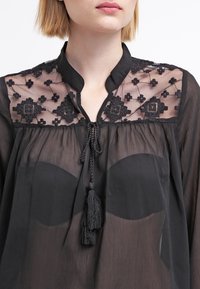 Zwarte doorzichtige blouse met een hoge halslijn, voorzien van geborduurde accenten op de kraag, een drawstring met franjes en een losse pasvorm.