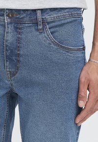 Blugi din denim într-o nuanță deschisă de albastru, cu cusături contrastante, închidere cu nasture și buzunare laterale. Suprafețele prezintă o ușoară estompare și textură.