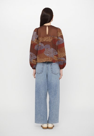 Brun mönstrad blus med puffärmar, med en nyckelhålsgång bak; kombinerad med vida blå denimjeans och ljusa skor.
