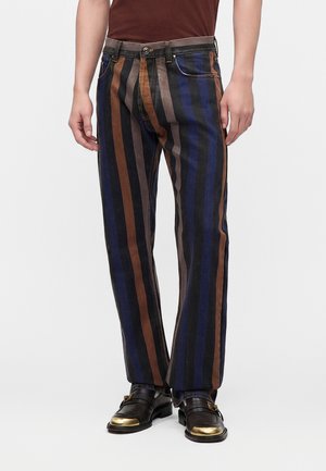 Homme portant un pantalon à rayures verticales marron, noir, gris et bleu, associé à des mocassins noirs avec des embouts dorés.