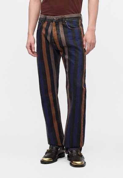Homme portant un pantalon à rayures verticales marron, noir, gris et bleu, associé à des mocassins noirs avec des embouts dorés.
