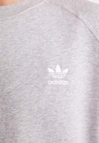 Szürke pulóver, puha textúrával, egy kis fehér hímzett logóval, amely három levelet és alatta az "adidas" szót tartalmazza.
