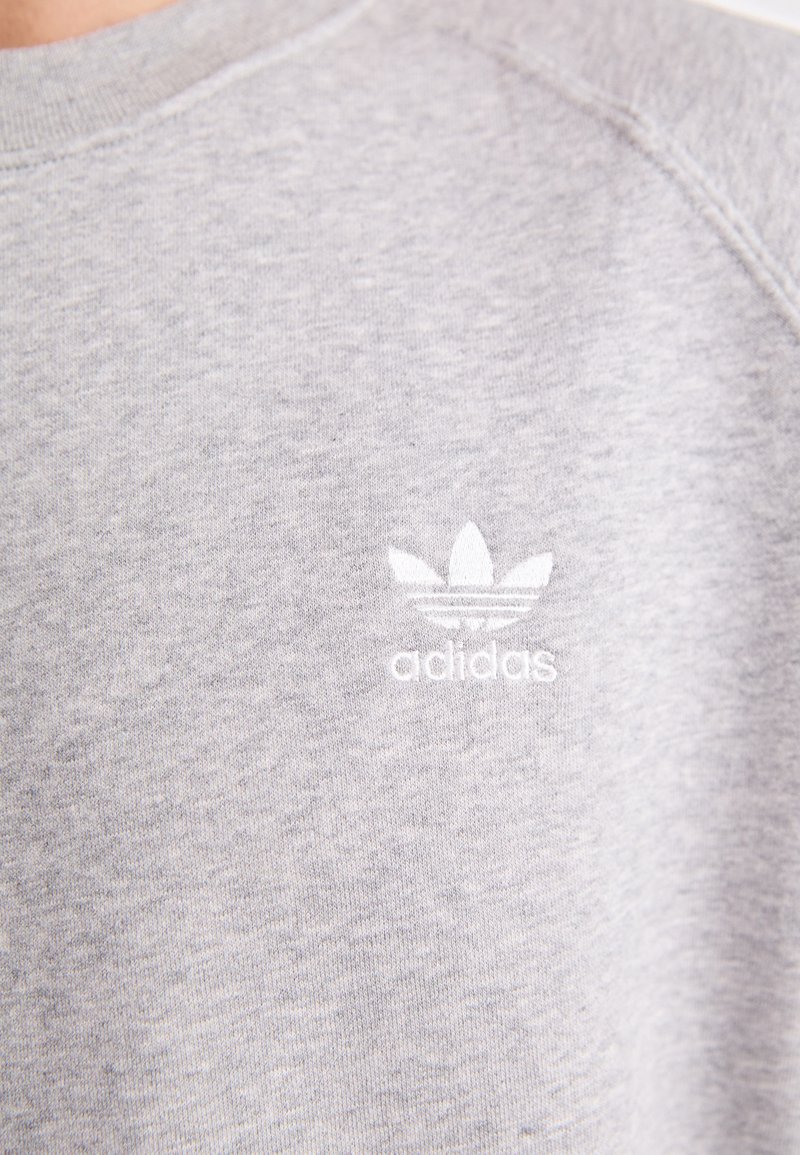 Sudadera gris con una textura suave, que presenta un pequeño logotipo bordado en blanco con tres hojas y la palabra "adidas" debajo.