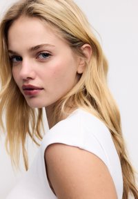 Jeune femme blonde aux cheveux longs et yeux bleus, portant un haut blanc sans manches, regardant légèrement par-dessus son épaule sur un fond uni.