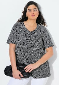 Černobílý top s paisley vzorem, véčkovým výstřihem, krátkými rukávy a volným střihem. Modelka drží černou kabelku a má na sobě bílé kalhoty.