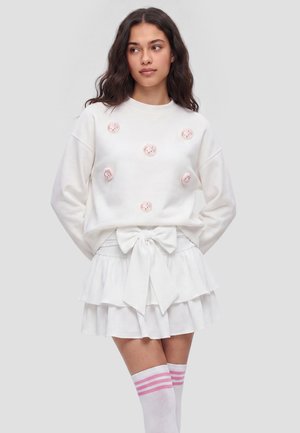 Ung kvinde med bølget hår iført en hvid sweatshirt med lyserøde tegneserieansigter, et hvidt skørt med en stor sløjfe og knæstrømper med lyserøde striber.