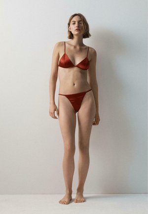 Kobieta stojąca boso przy jednolitej ścianie, ubrana w czerwony satynowy biustonosz i pasujące majtki, z krótkimi jasnobrązowymi włosami i neutralnym wyrazem twarzy.