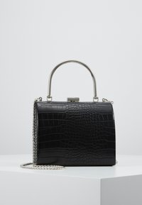 Musta suorakaiteen muotoinen käsilaukku krokotiilikuviolla, hopeanvärisellä metallikahvalla ja ketjusoljella, asetettuna valkoiselle pinnalle yksinkertaisen taustan edessä.