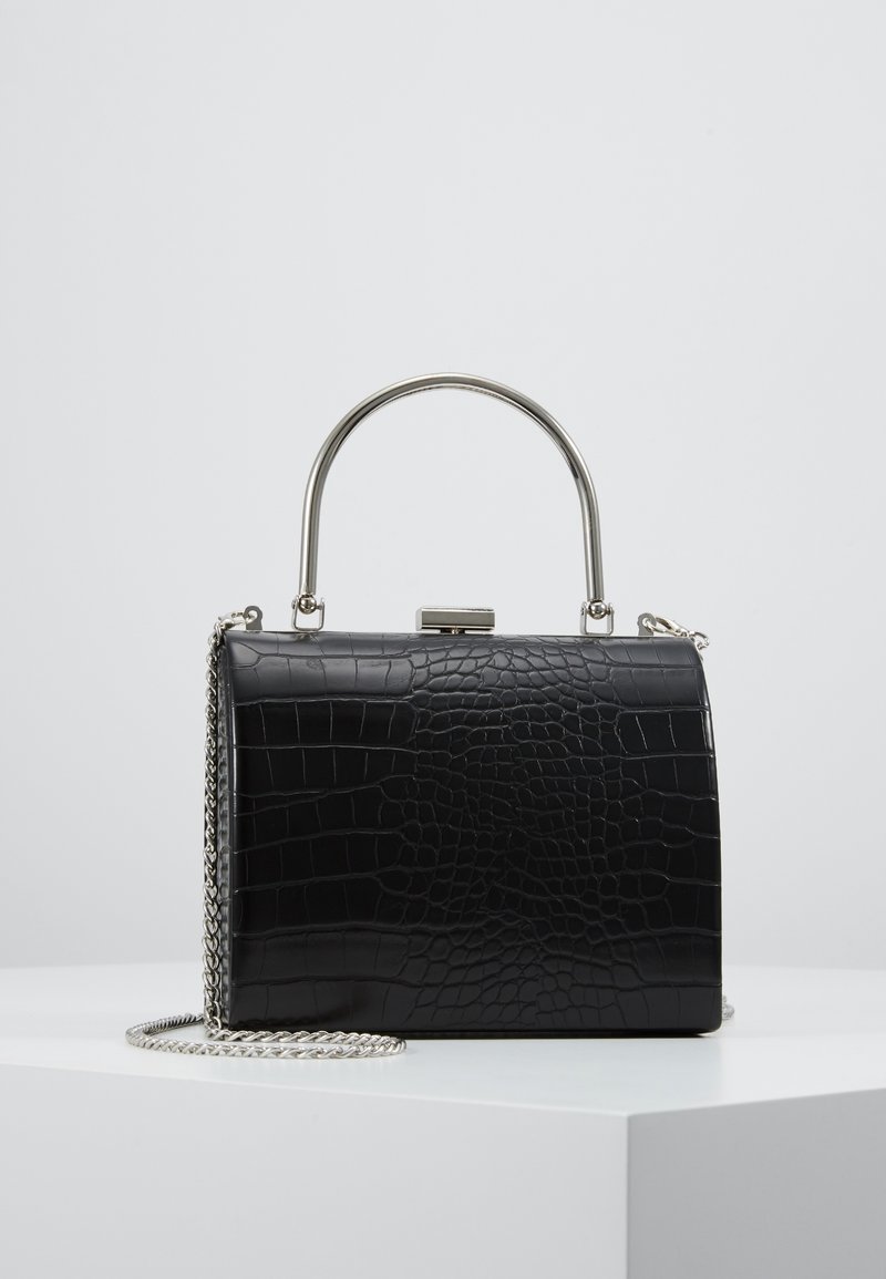 Musta suorakaiteen muotoinen käsilaukku krokotiilikuviolla, hopeanvärisellä metallikahvalla ja ketjusoljella, asetettuna valkoiselle pinnalle yksinkertaisen taustan edessä.