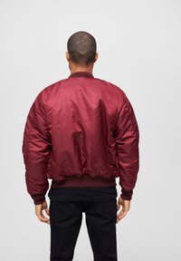 Veste aviateur bordeaux avec une texture lisse et brillante, des poignets et une taille côtelés ; présente des détails matelassés sur les manches. Pantalons noirs.