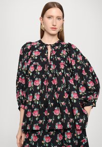 Svart blomstermønstrete bluse med rosa og grønne roser, rund hals, nøkkelhullsdetalj og puffermer. Myk stoffkvalitet, avslappet passform.