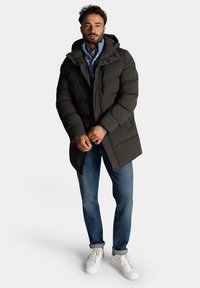 Donkergroene gevoerde parka met een capuchon, voorzien van een rits- en drukknoopsluiting, zijzakken en een gestructureerde afwerking. Gecombineerd met blauwe jeans en witte sneakers.