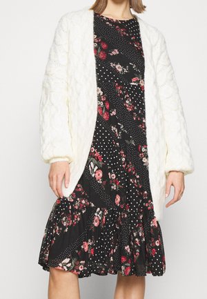 Kobieta w czarnej sukience w kwiaty i kropki, zestawionej z grubym, białym, dzianym kardiganem z balonowymi rękawami.