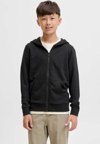Haut à capuche noir zippé avec des poches avant et des poignets côtelés. Porté par-dessus une chemise de couleur claire, associé à un pantalon beige.