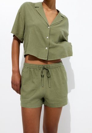 Femme portant une chemise courte boutonnée à manches courtes vert olive et un short à cordon assorti sur fond blanc.