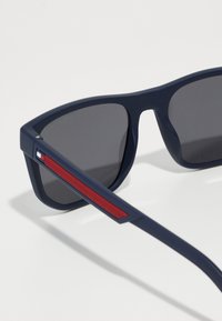 Navy blue sunglasses with gray lenses, featuring a textured red accent on the arm. The frame is angular and matte in finish.