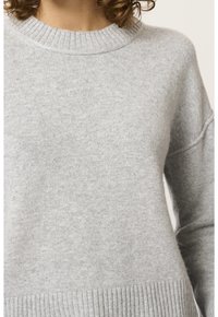 Femme portant un pull en maille gris clair à col rond avec col, poignets et ourlet côtelés, visible du cou jusqu'au milieu du torse.
