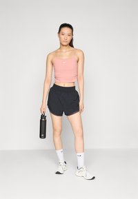 Idrettsantrekk bestående av en rosa croppet tank topp med hvit logo, sorte shorts, hvite sokker og sorte og hvite joggesko. Holder en svart vannflaske.