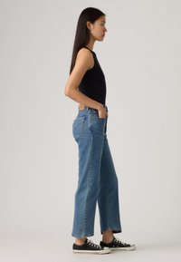 Μαύρο αμάνικο τοπ, μπλε τζιν με ίσια γραμμή ψηλόμεσο, χωρίς ραφή στο τελείωμα, και μαύρα πάνινα αθλητικά παπούτσια, φωτογραφημένα από το πλάι με φωτεινό φόντο.