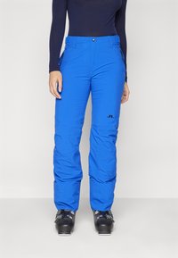 Μπλε μόνωσες σκι παντελόνες με ίσια γραμμή, τσέπες με φερμουάρ και μαύρη εμφάνιση λογοτύπου πάνω στο αριστερό μηρό. Το ύφασμα έχει λεία υφή.