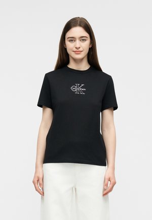 Młoda kobieta o długich brązowych włosach, ubrana w czarną koszulkę Calvin Klein i białe spodnie, stojąca na tle jednolitego jasnego tła.