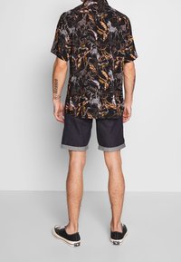 Man in een zwart T-shirt met korte mouwen en een kleurrijke dierenprint, donkere opgerolde shorts en zwarte sportschoenen, staand tegen een effen achtergrond.