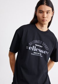 Svart bomull t-skjorte med rund hals, korte ermer, og et grafisk trykk i hvitt og grått som leser "ellesse" og andre tekstdetaljer.