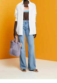 Chemise blanche boutonnée sur un top court multicolore, jeans évasés bleu clair, et un sac en toile lavande avec des accents en cuir. Fond orange.