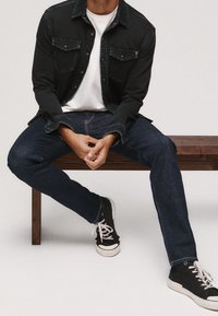 Svart denimjacka över en vit t-shirt, kombinerad med mörka denimjeans och svarta sneakers med vita sulor, sittande på en träbänk.