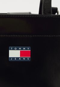 Sac à main en cuir verni noir avec un logo rectangulaire comportant des sections bleue, blanche et rouge, et le texte en relief "TOMMY JEANS" en dessous.