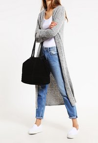 Lång, grå stickad cardigan med öppna sidor, bärs över en vit topp och blåjeans. Svart handväska och vita slip-on skor fulländar outfiten.