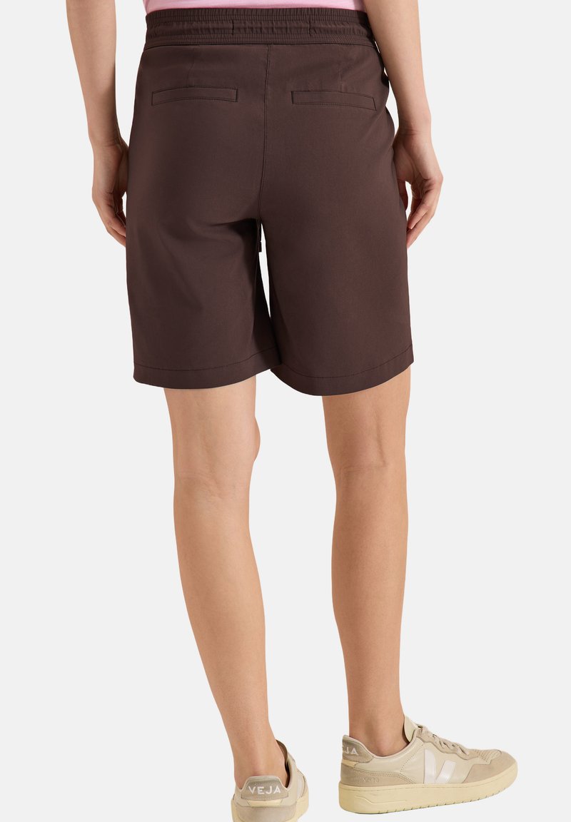 Personne portant un short marron foncé longueur genou avec des poches arrière et des baskets beiges, vue de dos sur fond uni.