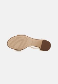 Beige sandaler med en slät, matt ovansidan och två remmar. Sulan är texturerad med spår och en brun gummiyta för grepp.