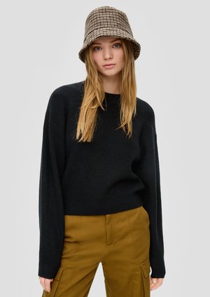 Sweater noir en maille côtelée avec épaules tombantes, associé à un pantalon cargo olive taille haute ; la personne porte également un chapeau seau à carreaux marron.