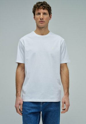 Hombre de pie mirando hacia adelante, vistiendo una camiseta blanca de manga corta lisa y jeans azules, contra un fondo gris liso.