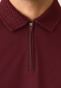 Vyno raudonio spalvos polo marškinėliai su užtrauktuku, puošiami tekstūruotu, raštuotu apykakliu ir lygia liemens dalimi, pagaminti iš minkštos medžiagos ir glaudžiai prigludę.