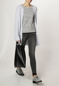 Helllila gestrickte Strickjacke über einem gestreiften schwarz-weißen Oberteil, schlanke dunkle Jeans, schwarze Tasche und gemusterte graue Schuhe.