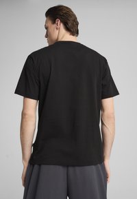Sort bomulds T-shirt med korte ærmer og rund hals; glat tekstur og afslappet pasform, set fra ryggen sammen med grå bukser.