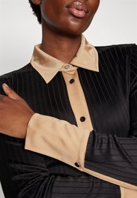 Personne portant une chemise noire côtelée avec un col, des poignets et une patte de boutonnage beige, les bras croisés sur la poitrine.