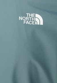 Logo The North Face v bílé barvě na světle modré látce s viditelnou texturou a jemnými záhyby.