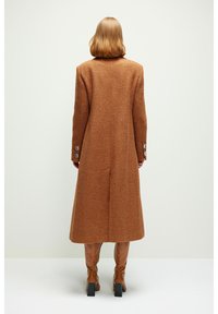 Femme aux cheveux mi-longs portant un long manteau marron texturé et des bottes assorties montant jusqu'aux genoux, debout devant un fond clair uni.