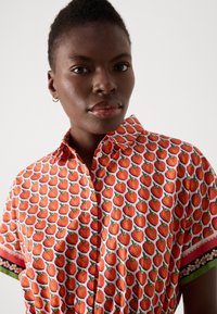 Kurzärmeliges Hemd mit einem roten Erdbeer-Muster auf weißem Hintergrund, mit gemustertem Saum an den Ärmeln und einem kragenförmigen Ausschnitt.