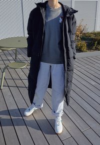 Schwarzer langer Daunenmantel, graues T-Shirt mit Grafik, hellgraue Jogginghose und weiße Sneakers mit grünen Akzenten, auf einem Holzdeck platziert.