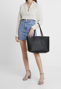 Mujer vestida con una camisa beige de botones y una falda de mezclilla, sosteniendo un gran bolso de cuero negro y usando sandalias de tacón cuadrado grises.