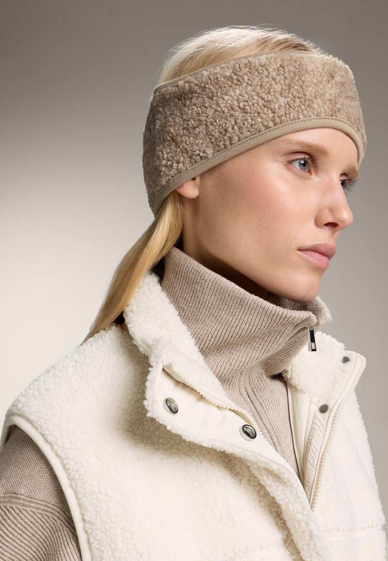 Beige fleece čelenka s měkkou texturou a kontrastním světle hnědým lemováním. Model má na sobě světle zbarvenou vestu a žebrovaný rolák.