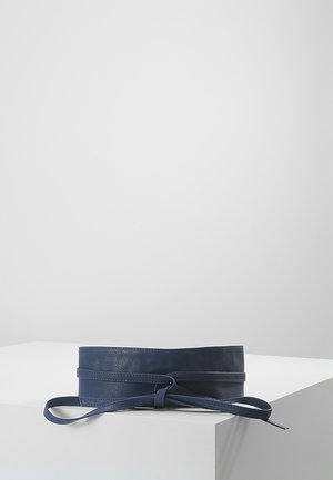 Ceinture de taille en cuir bleu marine avec une fine lanière nouée en nœud, exposée sur une surface blanche à angles, sur un fond gris clair uni.