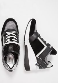 Baskets Michael Kors noires, blanches et argentées avec des lacets blancs, un tissu texturé et des accents métalliques au niveau du talon, présentées en vue de dessus et de côté.