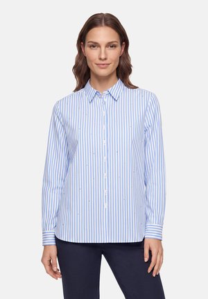Kvinde iført en lyseblå og hvid vertikalt striped knap-op skjorte med lange ærmer, stående mod en ensfarvet lys baggrund.