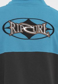 Blå og svart genser med brodert "Rip Curl"-logo i brunt og hvitt innenfor et ovalt design. Myk, teksturert stoff.